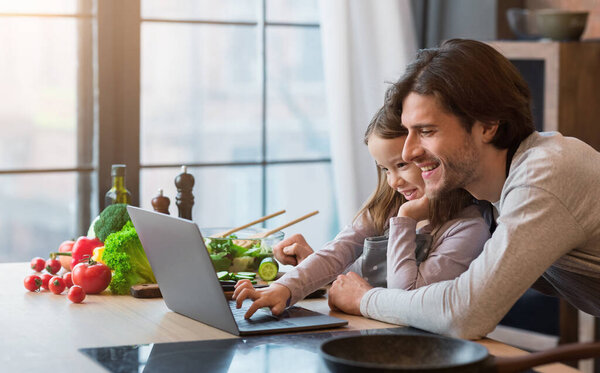Кулинарный блог. Веселая маленькая девочка и ее отец с помощью ноутбука на кухне
