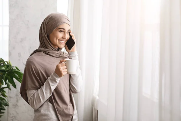Akhir pekan yang santai. Wanita Muslim meminum kopi dan berbicara di telepon seluler dekat jendela — Stok Foto