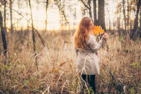 Portrait of cute redhead woman in coat holding yellow big leaf at autumn forest