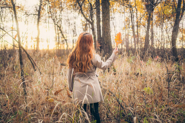 Portrait of cute redhead woman in coat holding yellow big leaf at autumn forest
