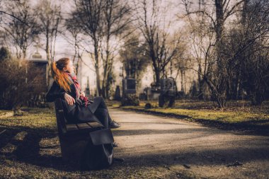 red hair female sitting on wooden bench at old cemetery path at Munich cemetery in Germany clipart