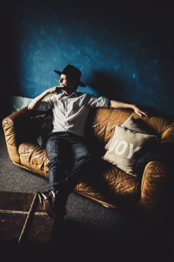 mavi duvar arka plan ile odasında kahverengi kanepede oturan şapkalı yakışıklı delikanlı 