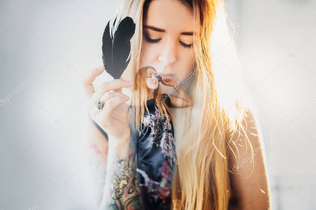 portrait of young fashion woman with closed eyes, red hair and black satin dress on room wall background holding black crow feather