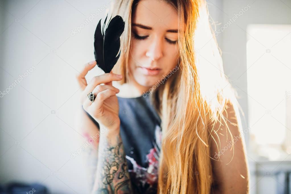 portrait of young fashion woman with closed eyes, red hair and black satin dress on room wall background holding black crow feather