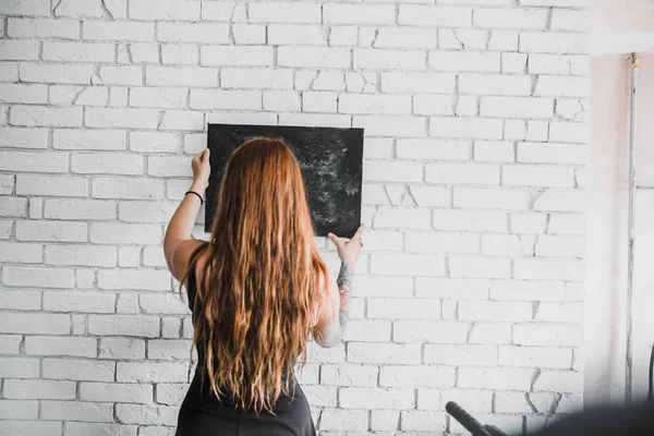 back view of young red hair woman in black dress holding black chalkboard on white brick wall background