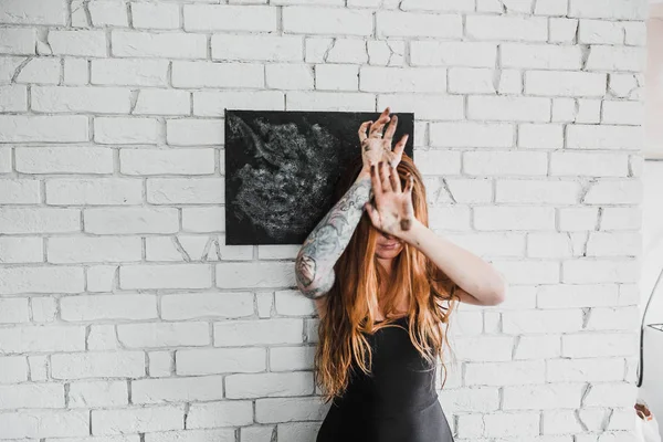 portrait of young red hair woman closed eyes by hands in black dress with black picture on white brick wall background