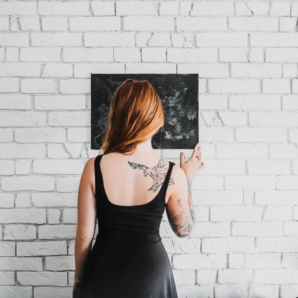 back view of young red hair woman in black dress holding black chalkboard on white brick wall background