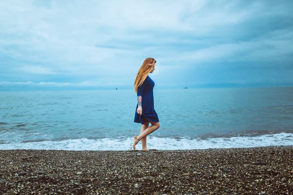 expression woman with long red hair in blue dress walking on stone beach at cloudy day outdoor