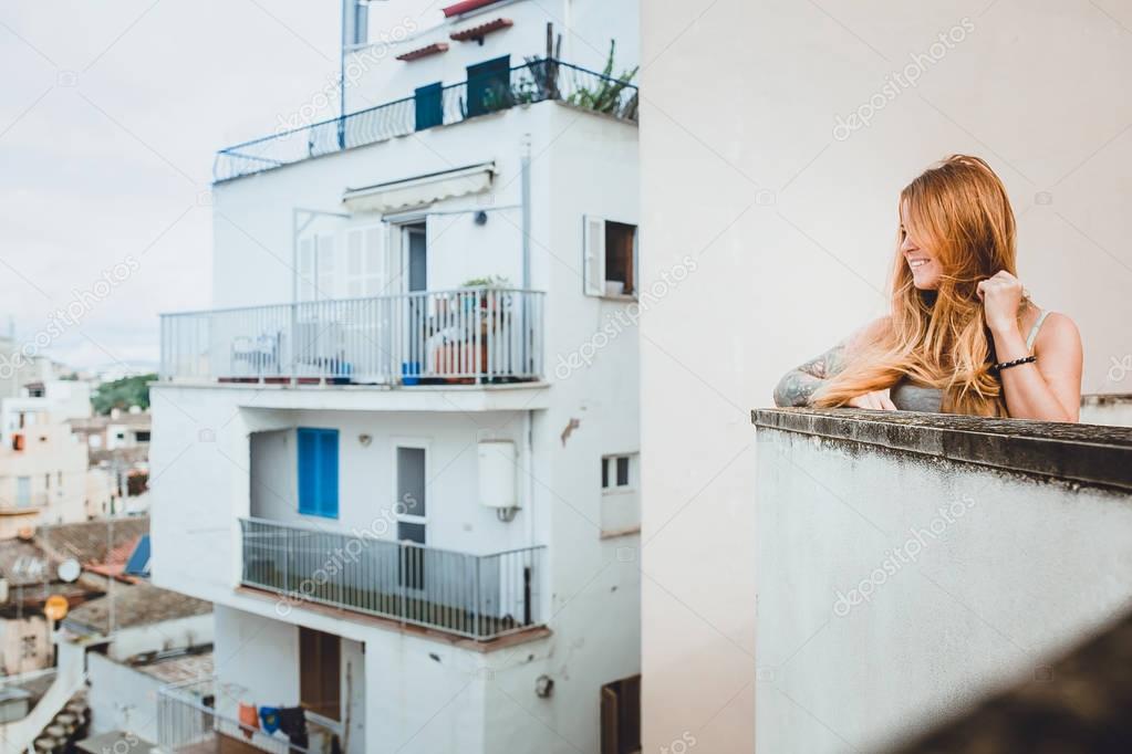 photo of cute young red hair woman with tattoo on hand on balcony of white building