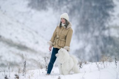 Genç bir kız kışın ormanda karlı bir çayırda beyaz Samoylu bir köpekle yürüyor..