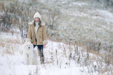 Genç bir kız kışın ormanda karlı bir çayırda beyaz Samoylu bir köpekle yürüyor..