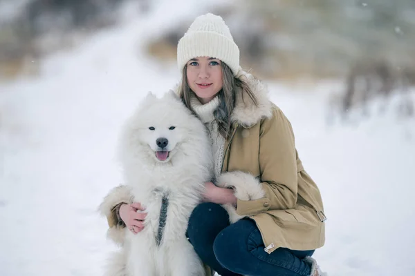 Genç bir kız kışın ormanda karlı bir çayırda beyaz Samoylu bir köpekle yürüyor..