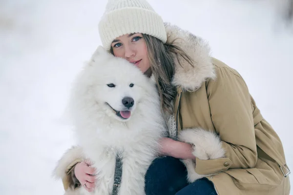 Genç bir kız kışın ormanda karlı bir çayırda beyaz Samoylu bir köpekle yürüyor..