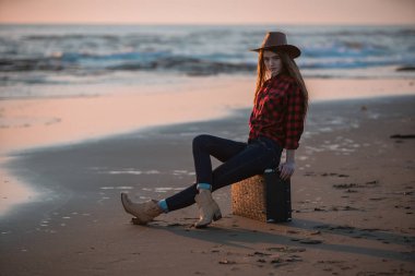 Kırmızı elbiseli, ince saçlı genç bir esmer gün batımında elinde bir bavulla, tropikal bir plajda çıplak ayakla denizin arka planında sörf yaparken yürüyor..