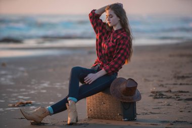 Kırmızı elbiseli, ince saçlı genç bir esmer gün batımında elinde bir bavulla, tropikal bir plajda çıplak ayakla denizin arka planında sörf yaparken yürüyor..