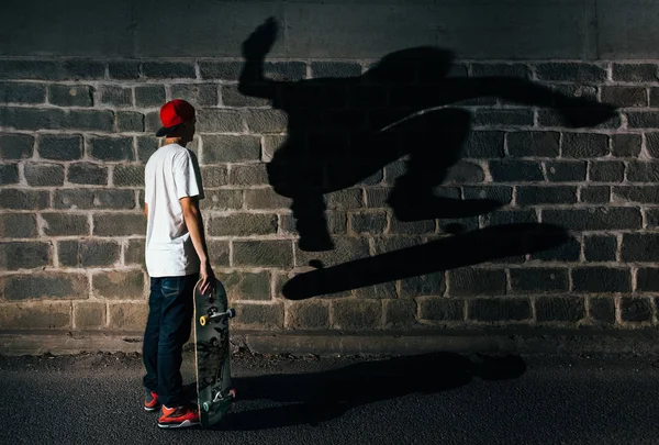 写真の左側に立って スタントの真ん中に自分の影を観察してカジュアルな服でスケートボーダー 影はアスファルトの道路の横の古い石の壁に描かれています — ストック写真