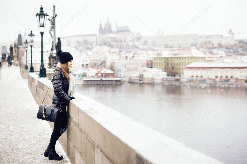 Woman is standing on bridge, leaning on brick railing. She is dressed in black winter clothing, wearing hat, gray scarf and black handbag over shoulder. City (Prague) in background is out of focus.