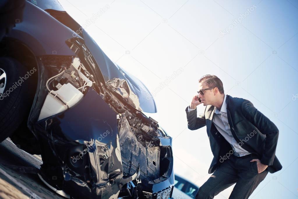 Male adult man in a suit and sunglasses checking damaged car while talking on the phone