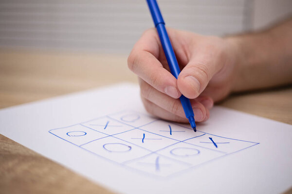 male adult hand holding blue pen above a paper with a tic-tac-to game