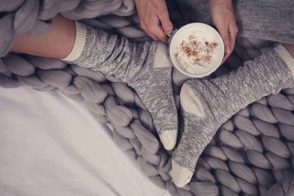 Женские ноги в уютных теплых шерстяных носках закрываются. Женщина, покрытая теплым одеялом, пьет кофе в постели  . — стоковое фото