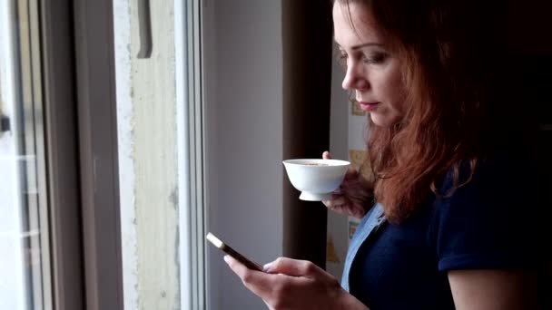 Молодая женщина пьет кофе с телефоном в руке и смотрит в окно во время карантина. Социальное дистанцирование — стоковое видео