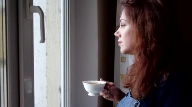 Karantina sırasında pencereden dışarı bakan genç bir kadın kahve içer. Sosyal uzaklık