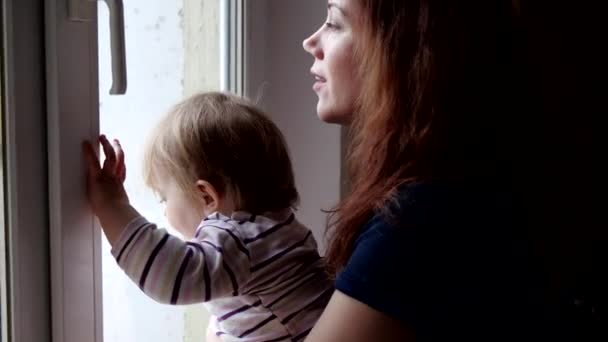 Una joven con un bebé mira por la ventana durante la cuarentena. distanciamiento social — Vídeos de Stock