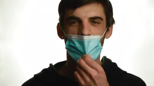 Een portret van een jonge man met een medisch masker die zijn masker afdoet en een pil neemt op een witte achtergrond — Stockvideo