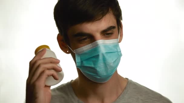 Retrato de un joven con una máscara médica que sacude una lata de pastillas y la mira sobre un fondo blanco — Vídeo de stock