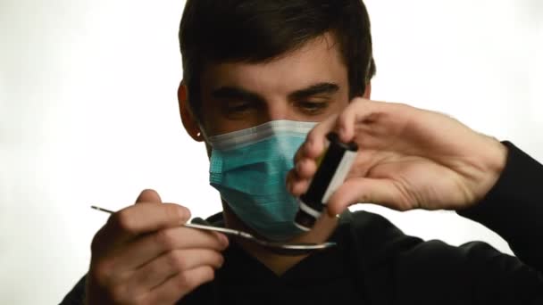 Ritratto di un giovane in una maschera medica rende gocce gocciolanti di medicina in un cucchiaio rimuove la maschera e bevande isolate — Video Stock