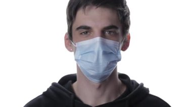 Tıbbi maskeli bir gencin portresi termometreye bakar ve beyaz arka plandaki alnın sıcaklığını kontrol eder.