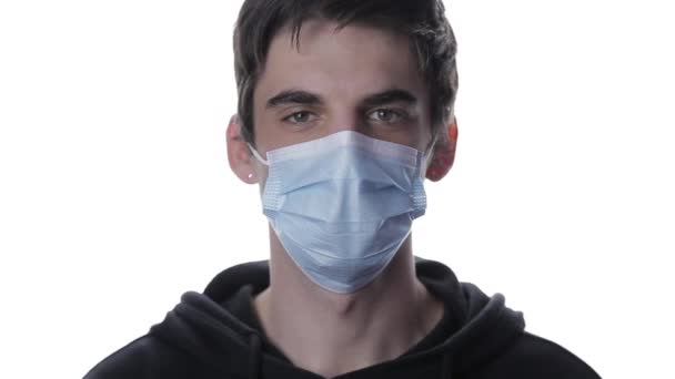 Ett porträtt av en ung man i medicinsk mask tittar på termometern och kontrollerar temperaturen i pannan på en vit bakgrund — Stockvideo