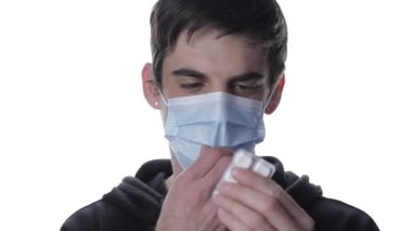 Maskesini çıkarıp beyaz arka planda hap alan tıbbi maskeli bir gencin portresi.