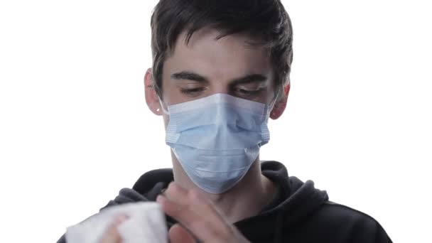 Tıbbi maskeli genç bir adamın portresi beyaz arka planda antibakteriyel peçeteyle ellerini siliyor. — Stok video