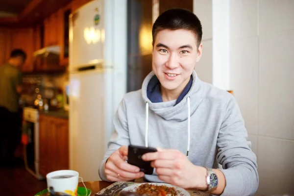 Sentimiento positivo de un joven sentado en la cocina durante el desayuno sostiene en sus manos el teléfono, mirando a la cámara . — Foto de Stock