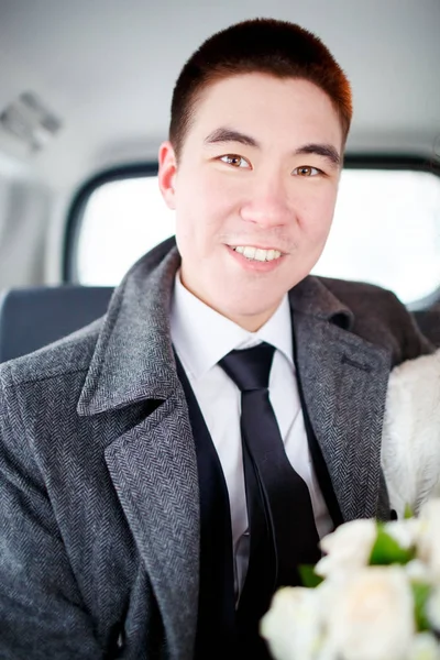 El novio en un abrigo de invierno sentado en el coche con ramo de bodas en las manos. Retrato positivo, mirando a la cámara . — Foto de Stock