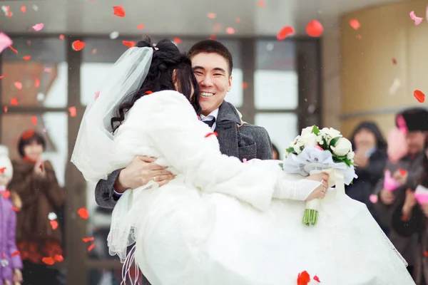 El novio llevando a la novia en sus brazos, la multitud lanza pétalos y arroz. Feliz boda. . Fotos De Stock