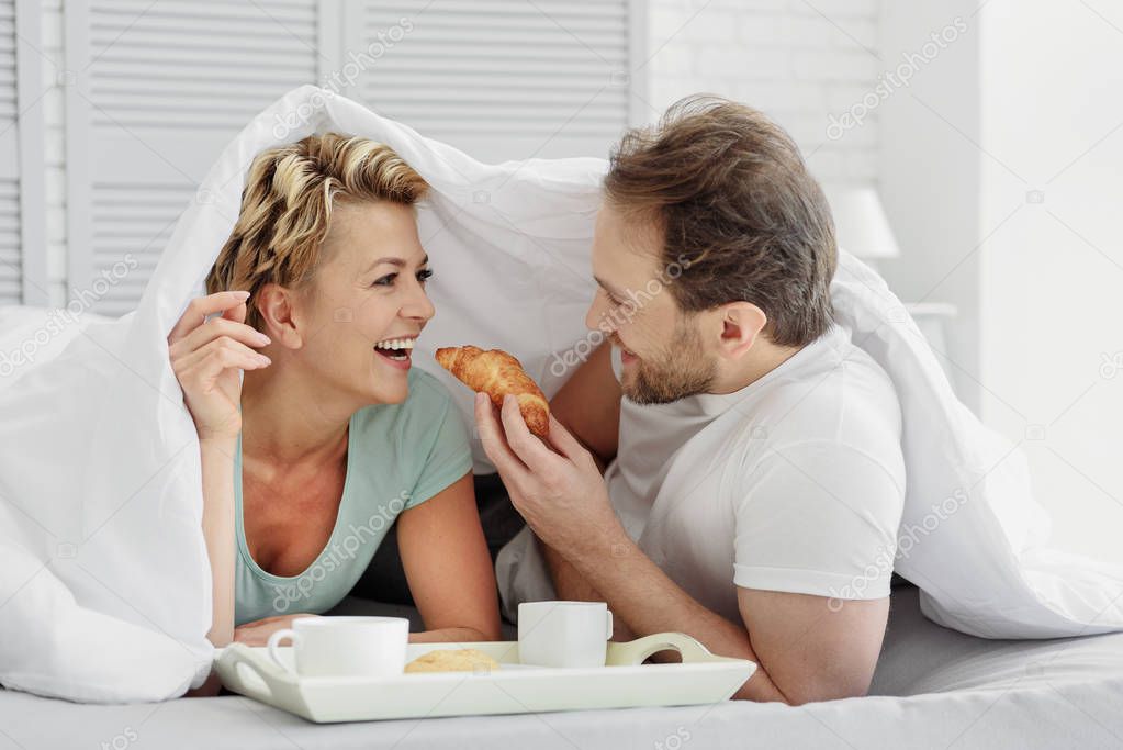 Joyful married couple eating breakfast in bedroom