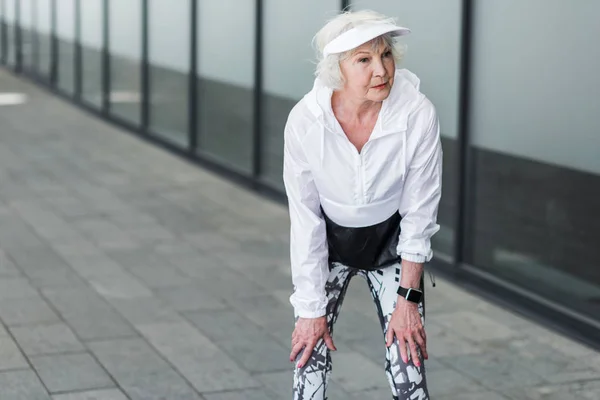 Грустная пожилая женщина отдыхает после физических упражнений возле здания города — стоковое фото