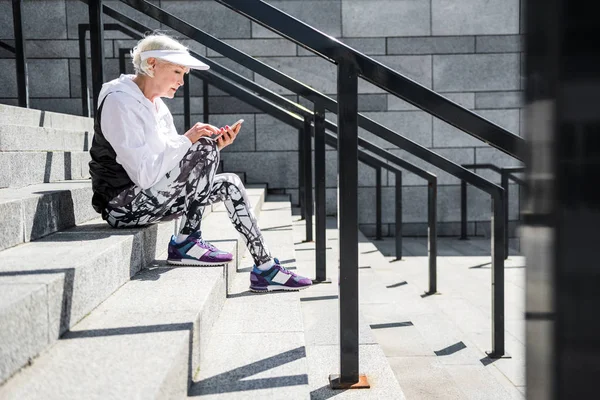 Концентрированная пожилая леди, которая пишет через свой мобильный телефон на каменных ступеньках — стоковое фото