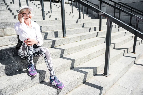 Радостная пожилая женщина с помощью смартфона, опираясь на ступени структуры — стоковое фото