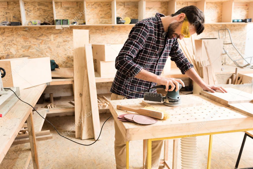 Cheerful woodworker is leaning on table while laboring with instrument