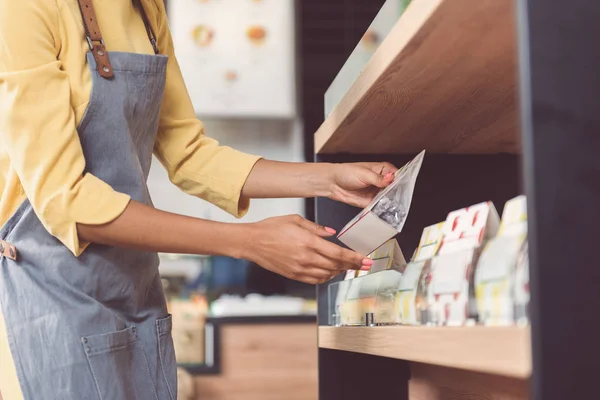 Pleasant girl in apron is putting new products on rack — Stock Photo, Image