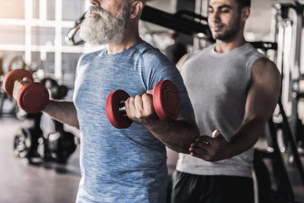 Old bearded man having workout in gym with professional instructor