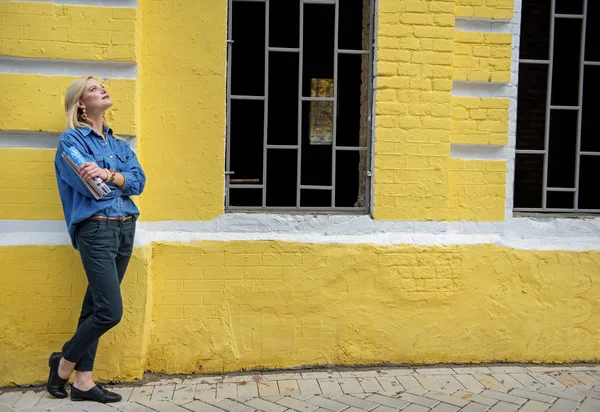 Dreamful blond girl standing near yellow building — Stock Photo, Image