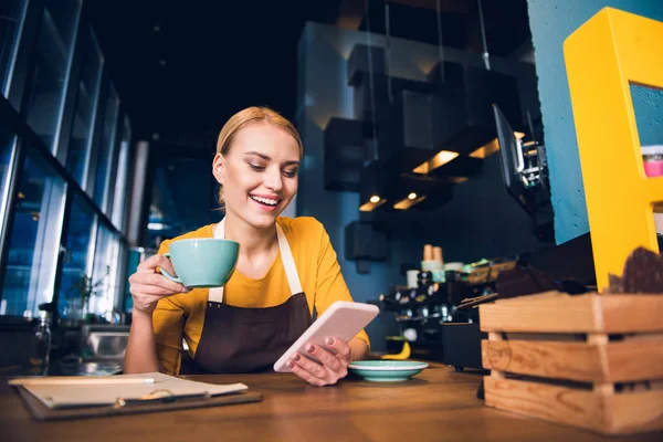 Cheerful girl barista typing in phone