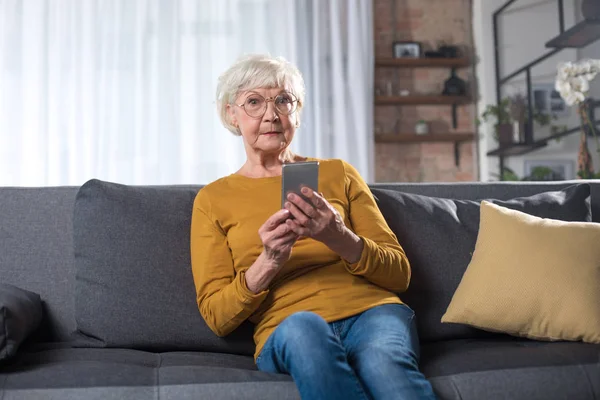 Вражена стара леді відпочиває на дивані у вітальні з телефоном — стокове фото