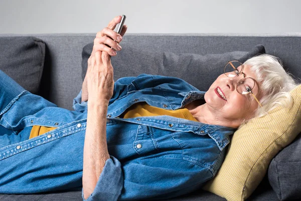 Anciana alegre tumbada en un sofá con teléfono móvil — Foto de Stock