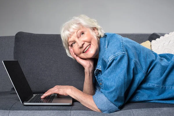 微笑的老妇人躺在舒适的沙发上与笔记本 — 图库照片