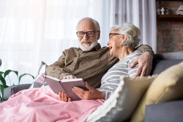 Encantada pareja de ancianos sentados en un sofá con libro —  Fotos de Stock